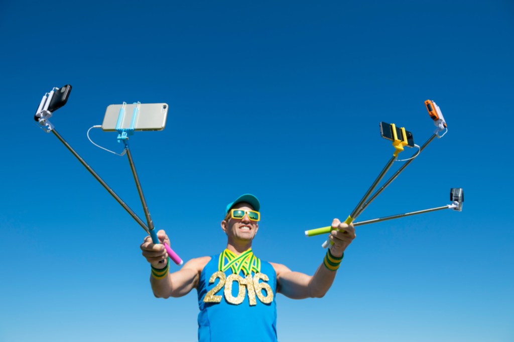 A man wearing sunglasses and a blue tank top with '2016' printed on it is holding three selfie sticks, each with a smartphone attached, against a clear blue sky.